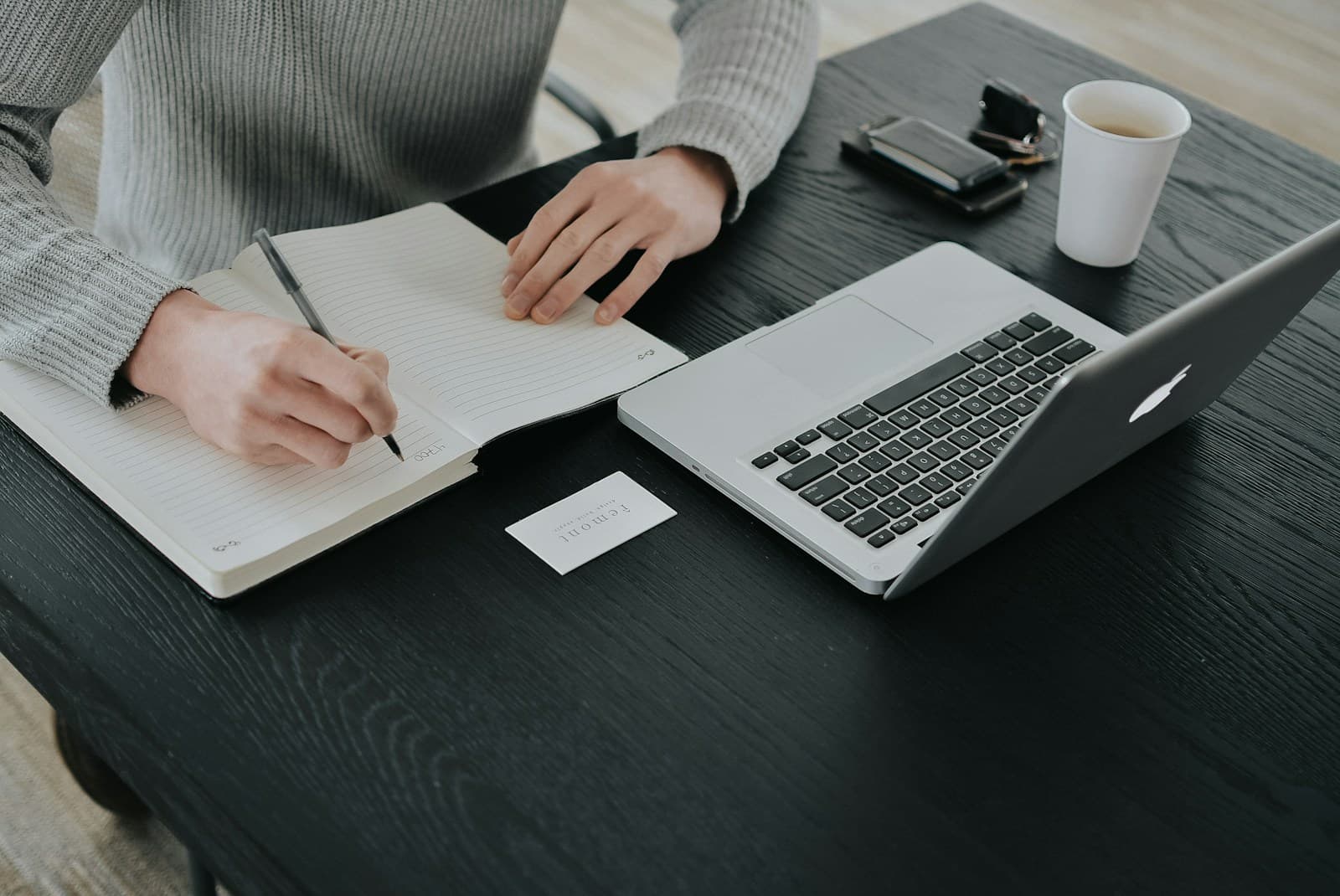 Person in a gray sweater writing in a notebook beside a laptop and coffee cup